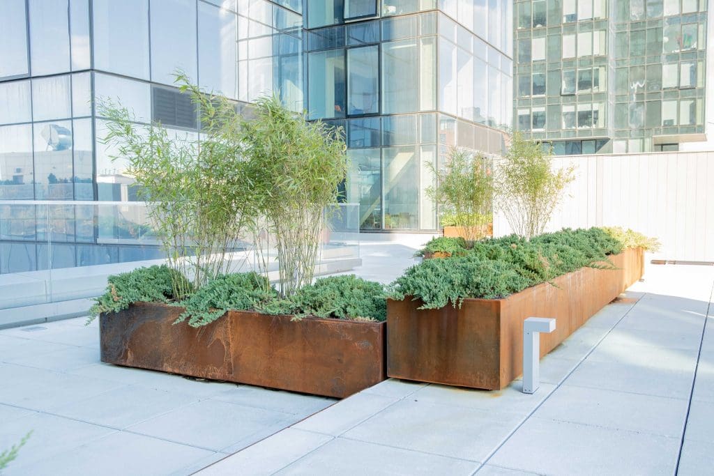A number of Corten Steel Planters on a stone walkway arranged in a L-shape